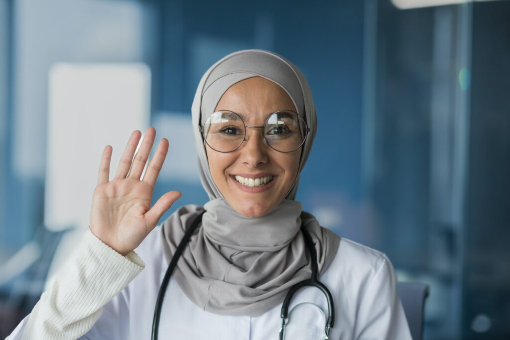 A woman, a Muslim, Arab woman doctor. She stands dressed in a hijab, glasses and with a stethoscope